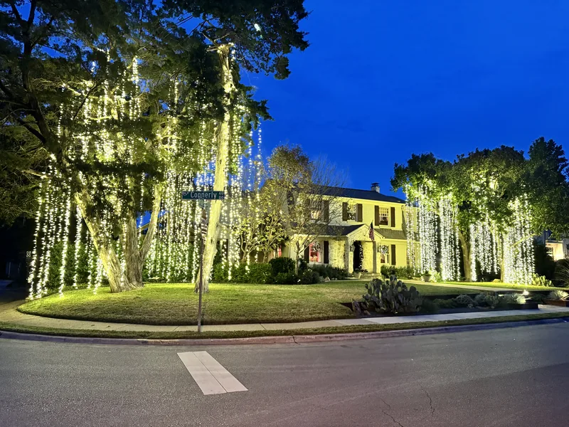 Christmas light installation by Christmas Lights Experts DFW &mdash; mature trees draped in cascading warm white lights in front of a white two-story home